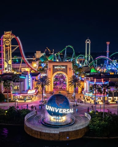 Nighttime aerial view of theme park with illuminated globe, roller coasters, castle, and colorful rides reflected in water