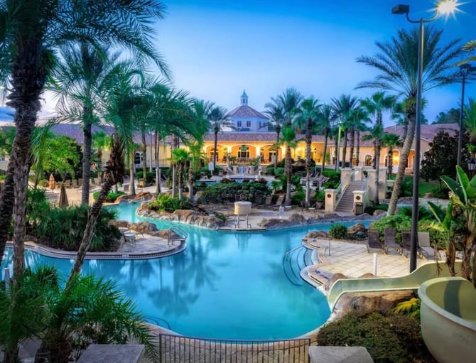 Resort pool complex with palm trees, illuminated yellow building with cupola, and tropical landscaping at dusk